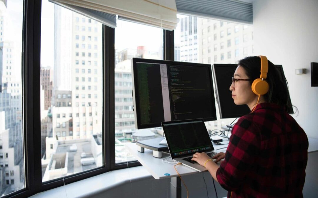 A developer sits at their desk in front of two big monitors, looking out of the window at a city skyline.