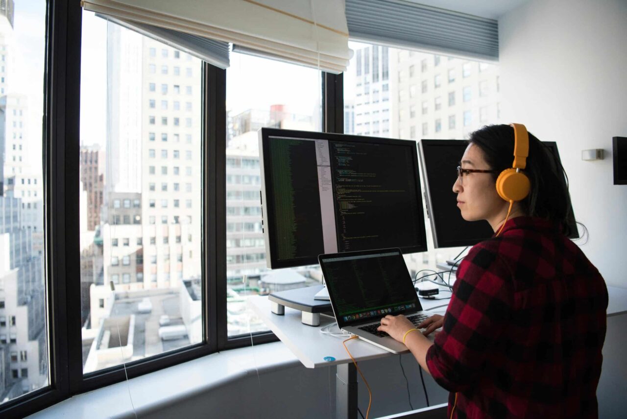 A developer sits at their desk in front of two big monitors, looking out of the window at a city skyline.