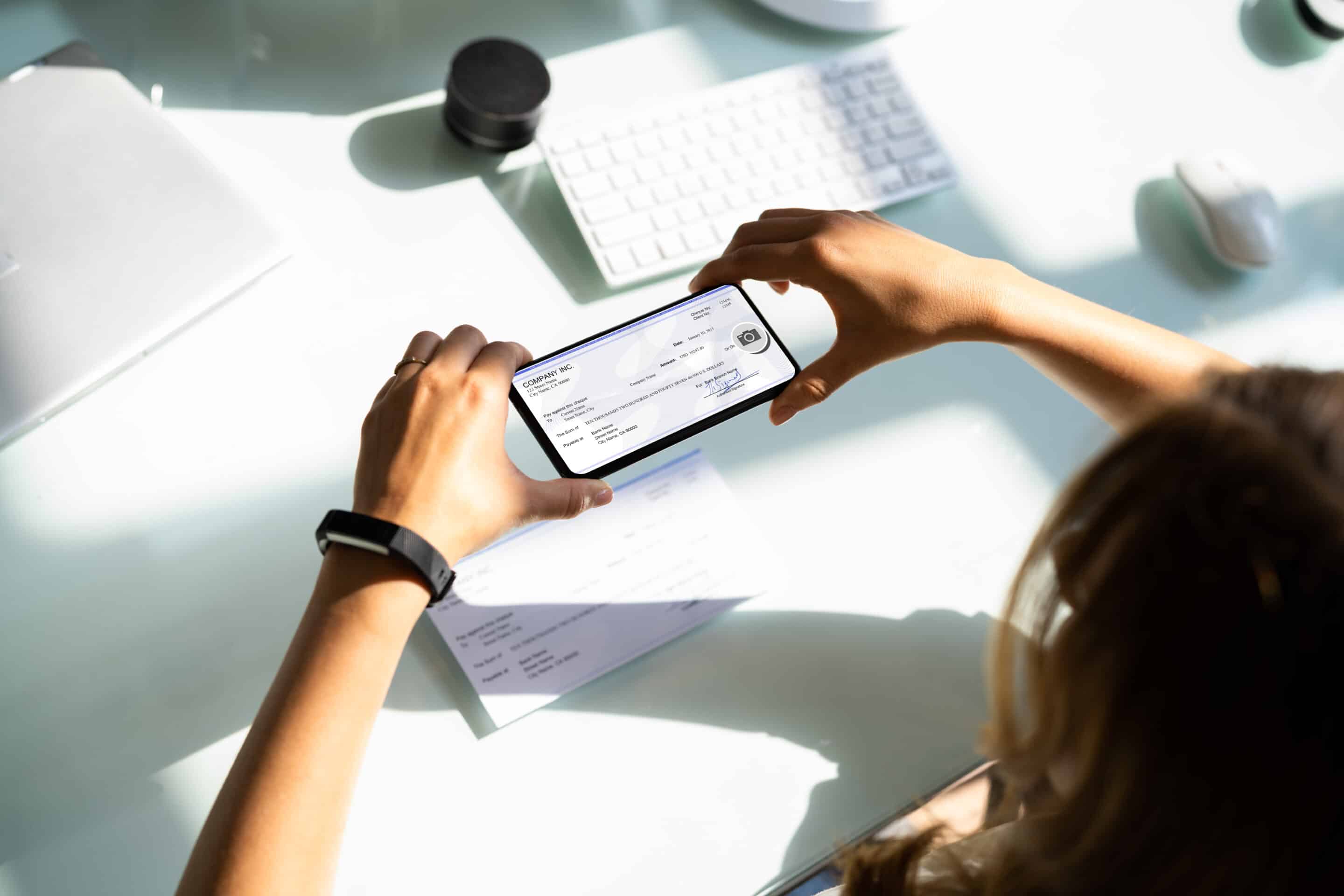 A woman's hands holding a mobile phone over a check on a white desk. The phone screen shows an image of the check. A keyboard and coffee mug are also on the desk.
