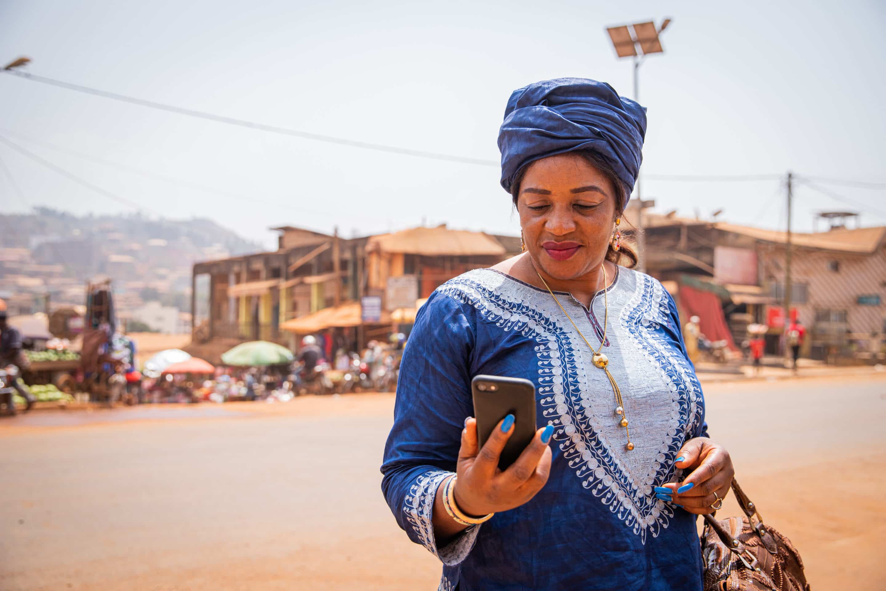 An African woman dressed in a traditional dress looks at the screen of her smartphone. 50-year-old woman in an African village uses technology A woman in African attire holding a mobile device