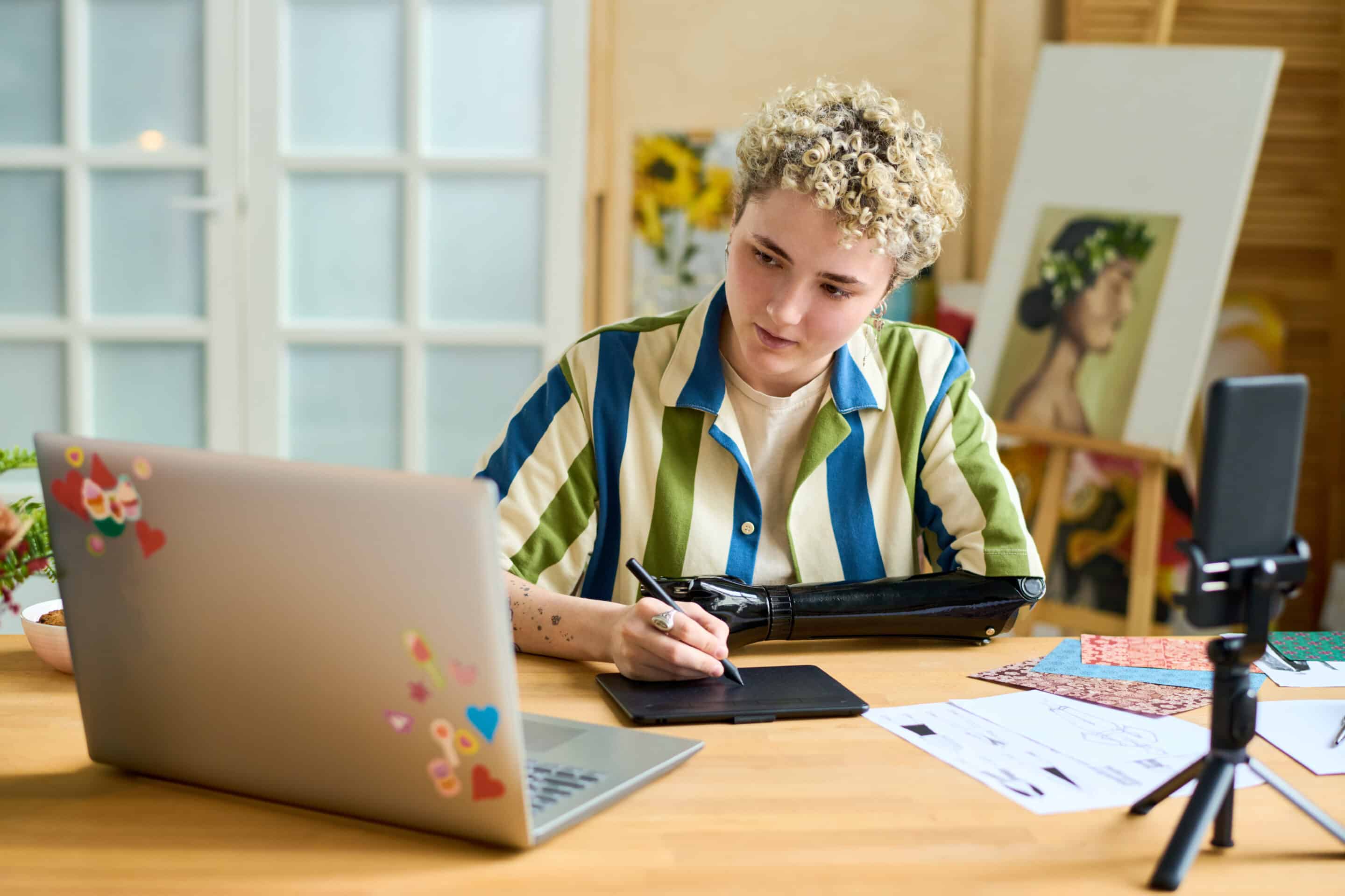 A young graphic designer with an arm prosthesis creating a new drawing while looking at laptop screen in front of smartphone camera