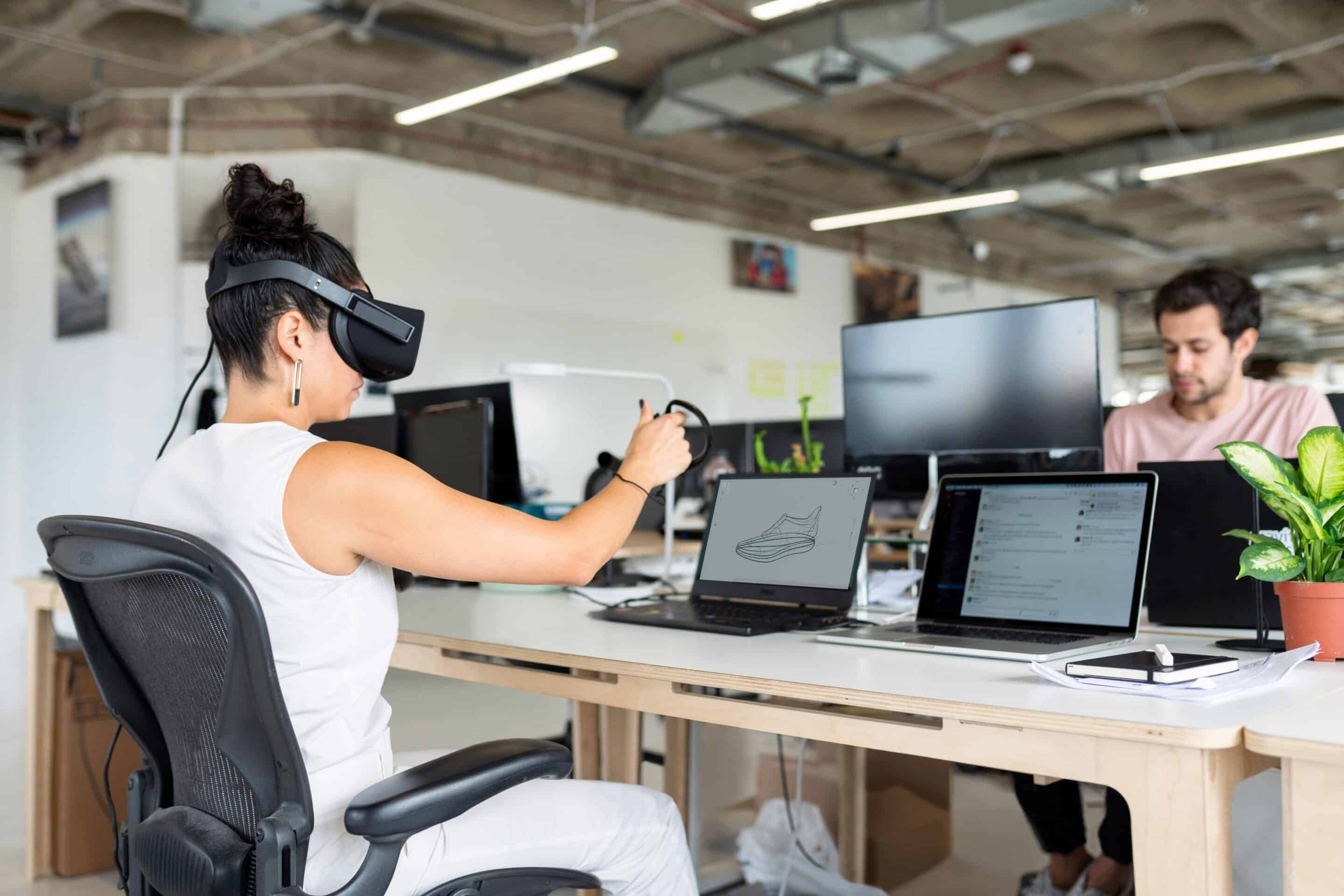 A man sits at a desk with several computers. Across from him, a woman sits wearing a VR headset in front of two laptops.