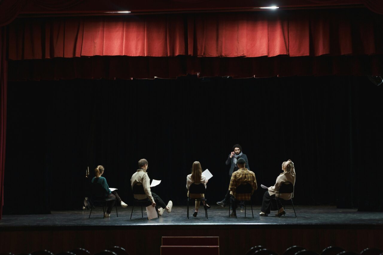Performers sit and read scripts on a stage.