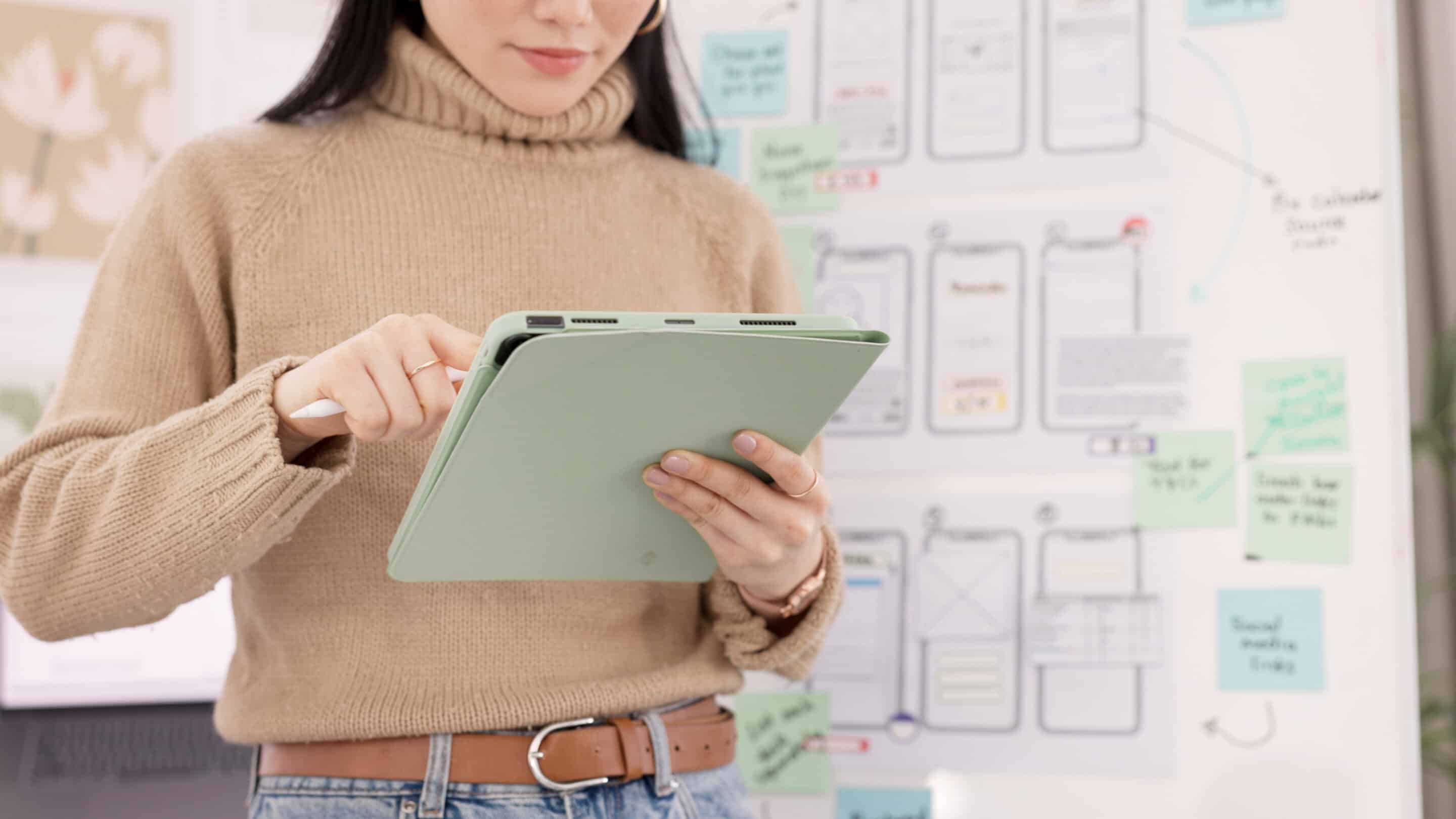 Young woman with shoulder-length brown hair, beige sweater holding tablet in front of whiteboard being used for UX design.