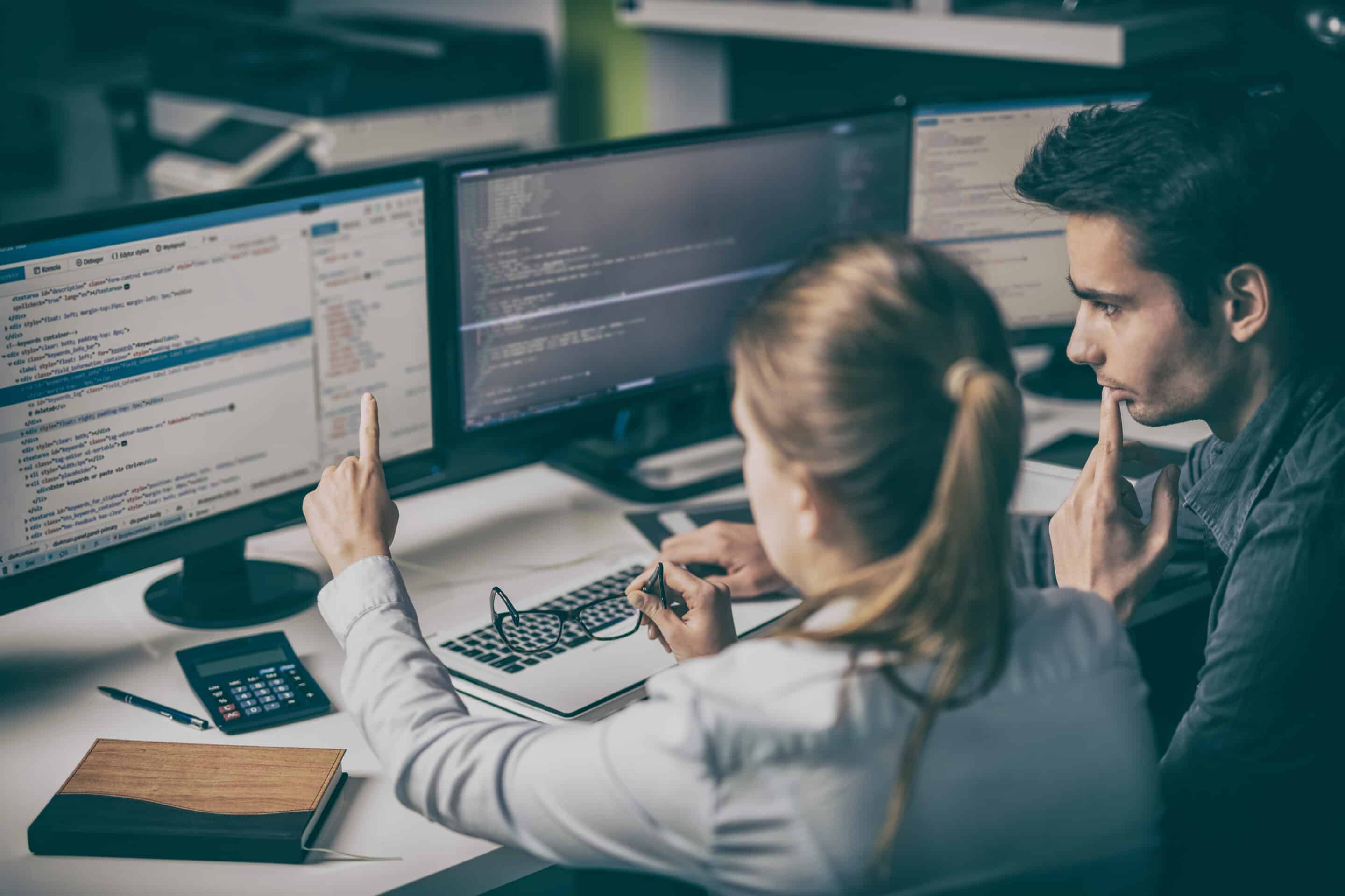 Woman web designer with blonde hair points at web design screen while male colleague with dark hair looks on.