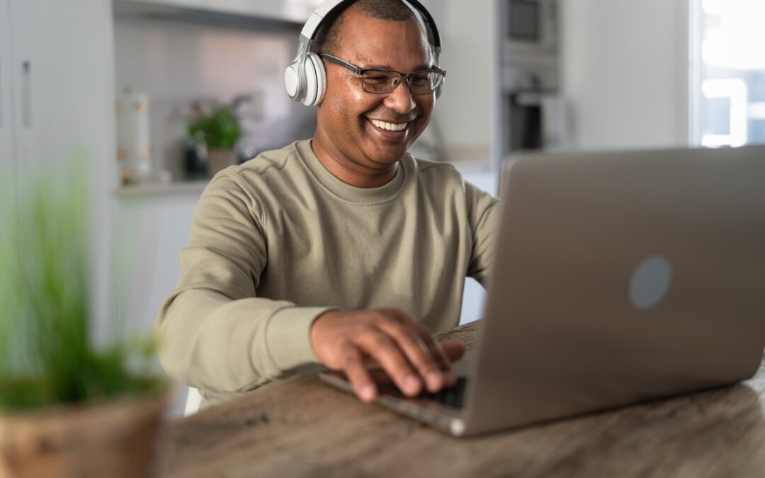 A man smiles while wearing headphones and typing on his laptop keyboard.