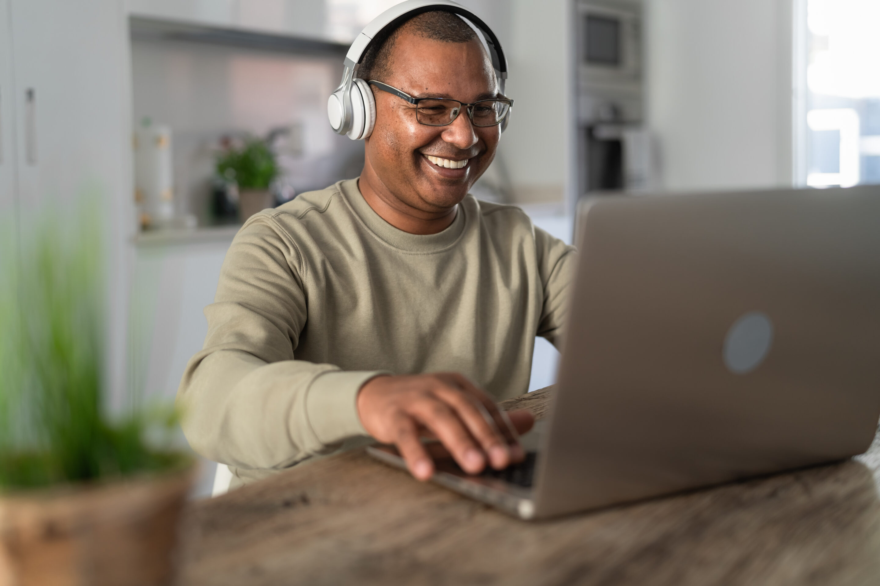 A man smiles while wearing headphones and typing on his laptop keyboard.