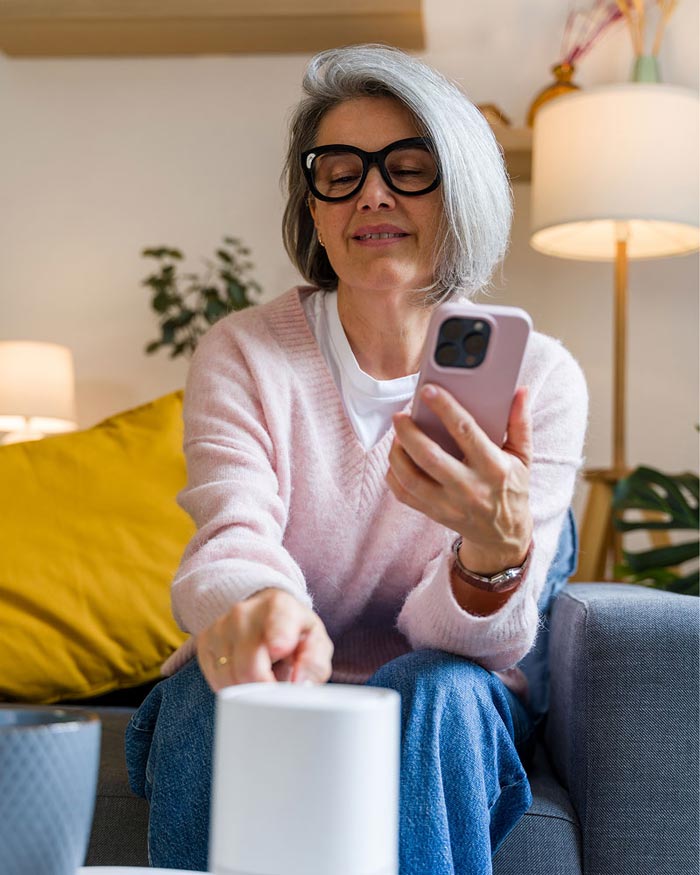 ai-data-section-1 An older woman with glasses holding a smartphone next to a smart home speaker in her living room.