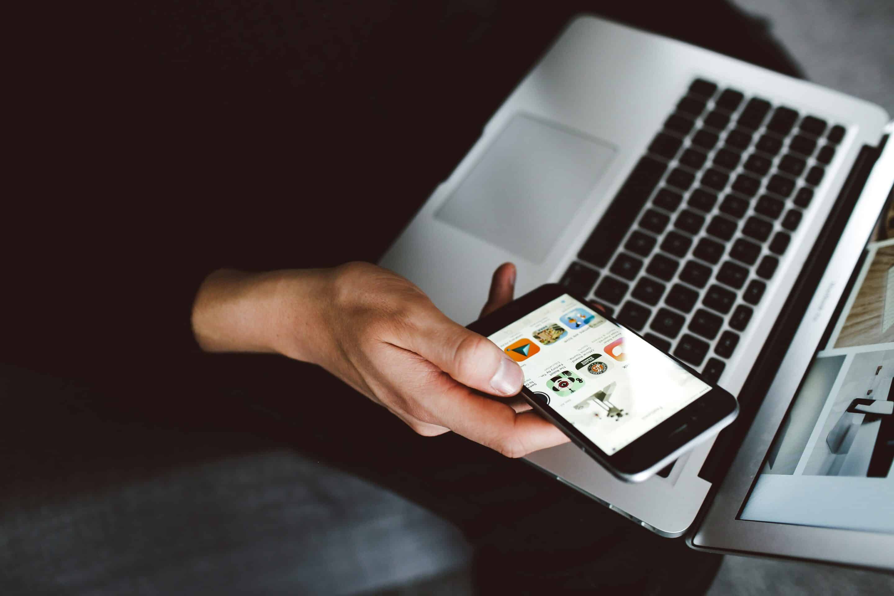 A close-up of a hand holding a mobile phone appears slightly above an laptop.
