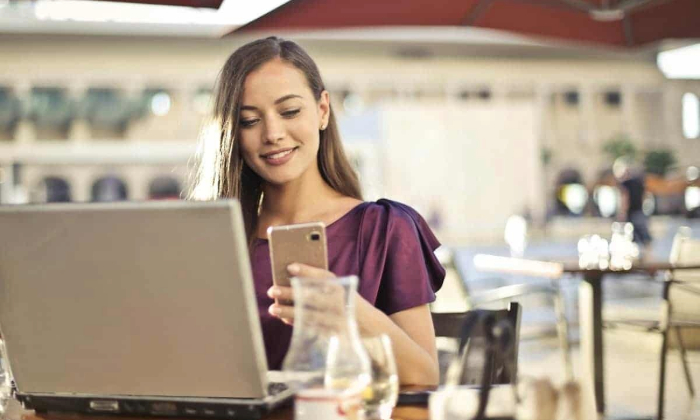 A woman uses a laptop while looking at her phone.