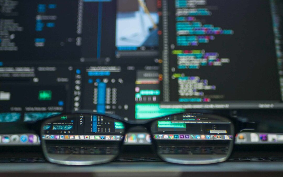 A pair of glasses sits at a desk in front of a monitor filled with many applications open.